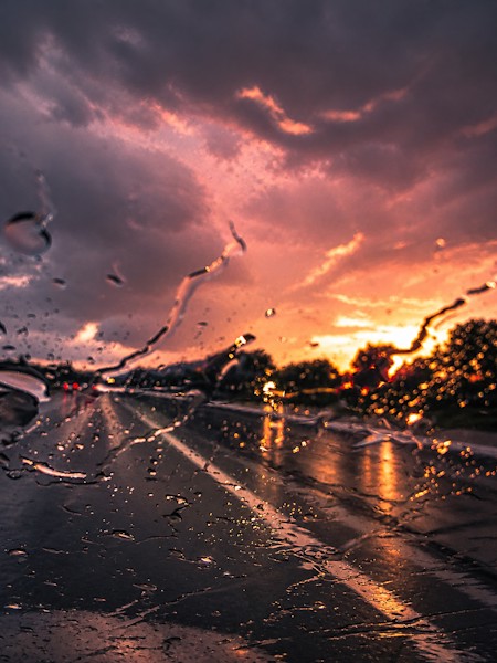 A dramatic sunset viewed through a rain-covered windshield, with glowing reflections on the wet road and warm golden light beneath a moody purple sky. A atmospheric scene capturing the beauty of rain, light, and evening weather.